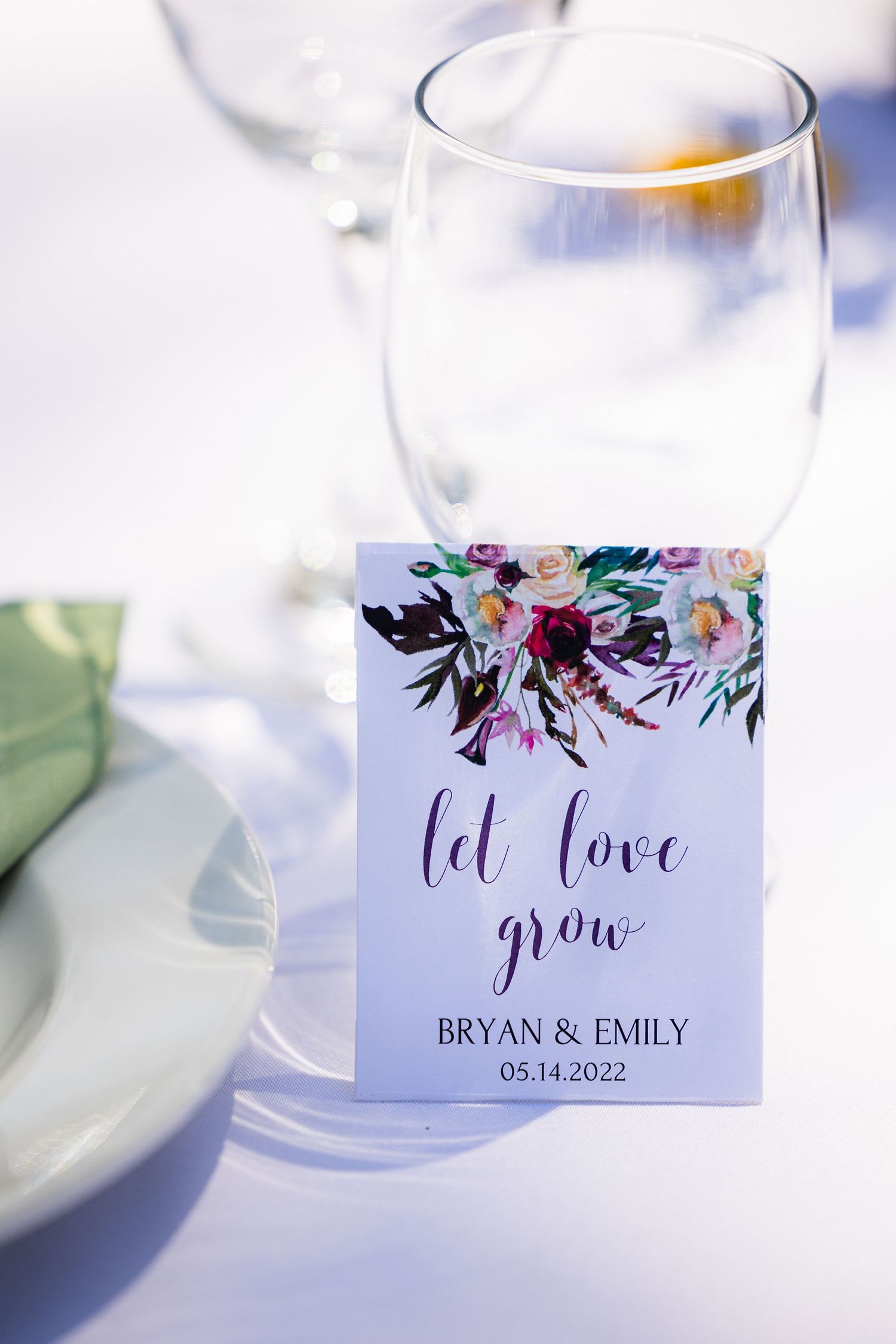 Wedding favor tag with floral design and 'let love grow' text next to empty wine glass on white tablecloth.