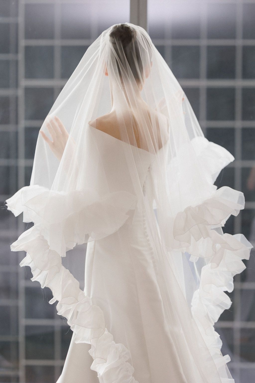 Back view of a bride wearing an elegant white dress with a dramatic ruffled veil against window light.