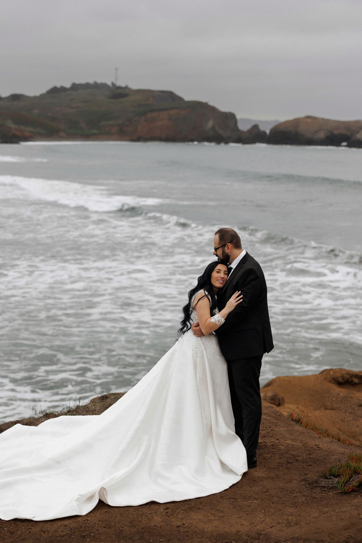 A couple shares an intimate moment on a dramatic coastal cliff overlooking the ocean during their wedding photoshoot.