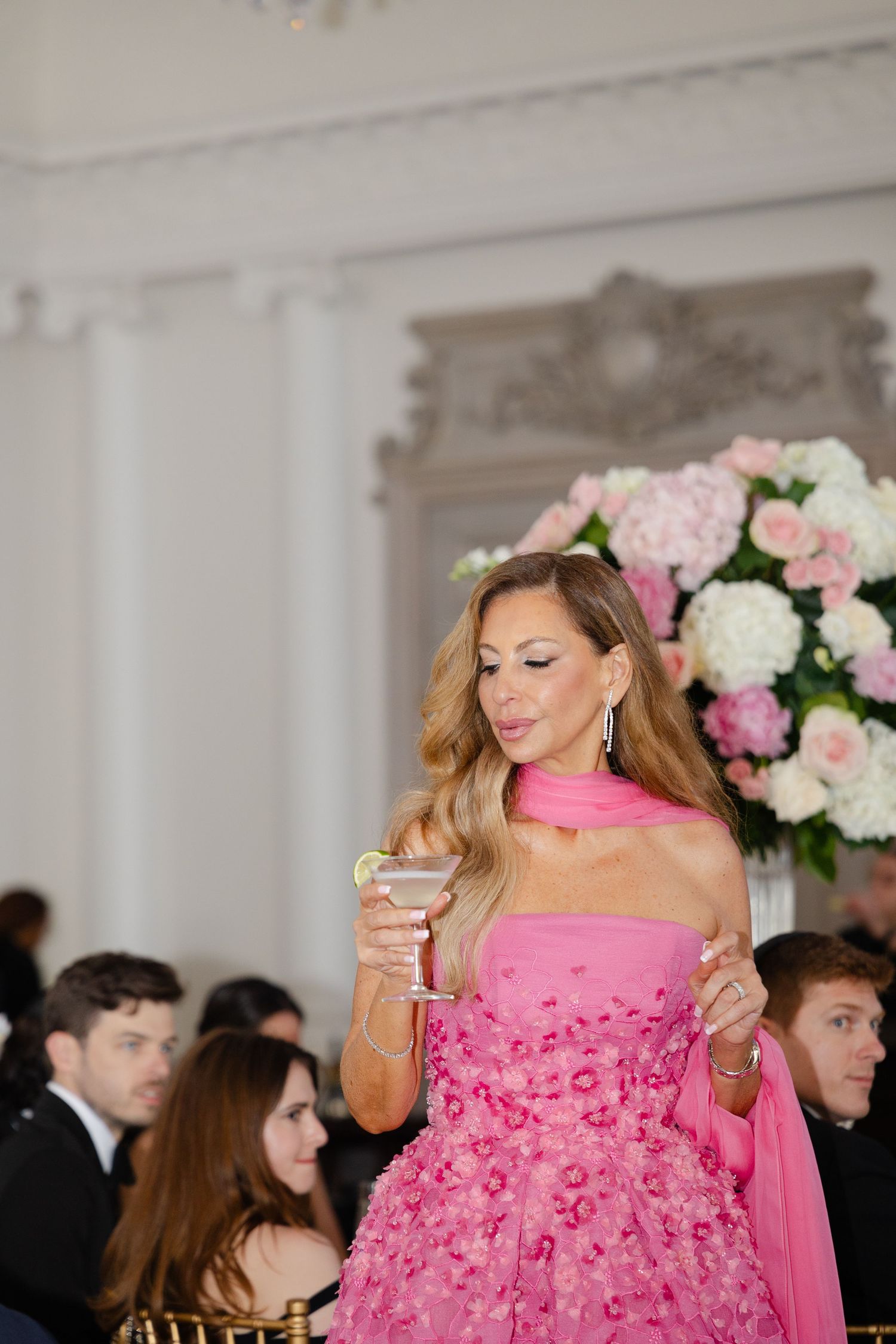 A person in a vibrant pink ruffled gown holds a champagne glass at an elegant event with floral decorations.