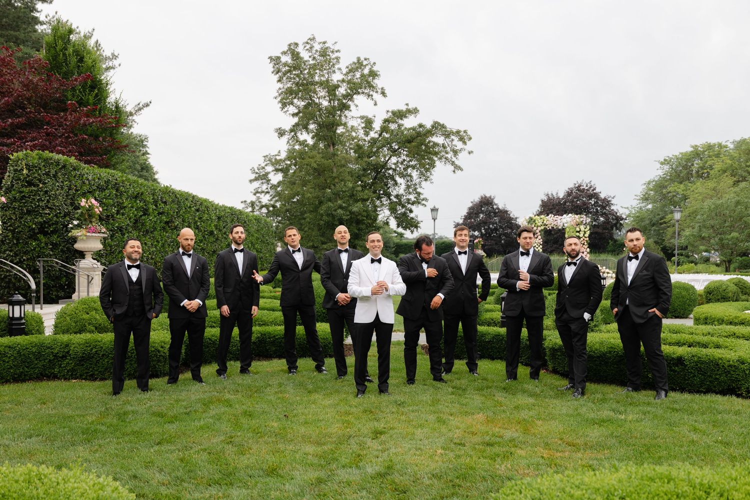 A group in black tuxedos and one person in white stands together on landscaped grounds for a formal event photo.