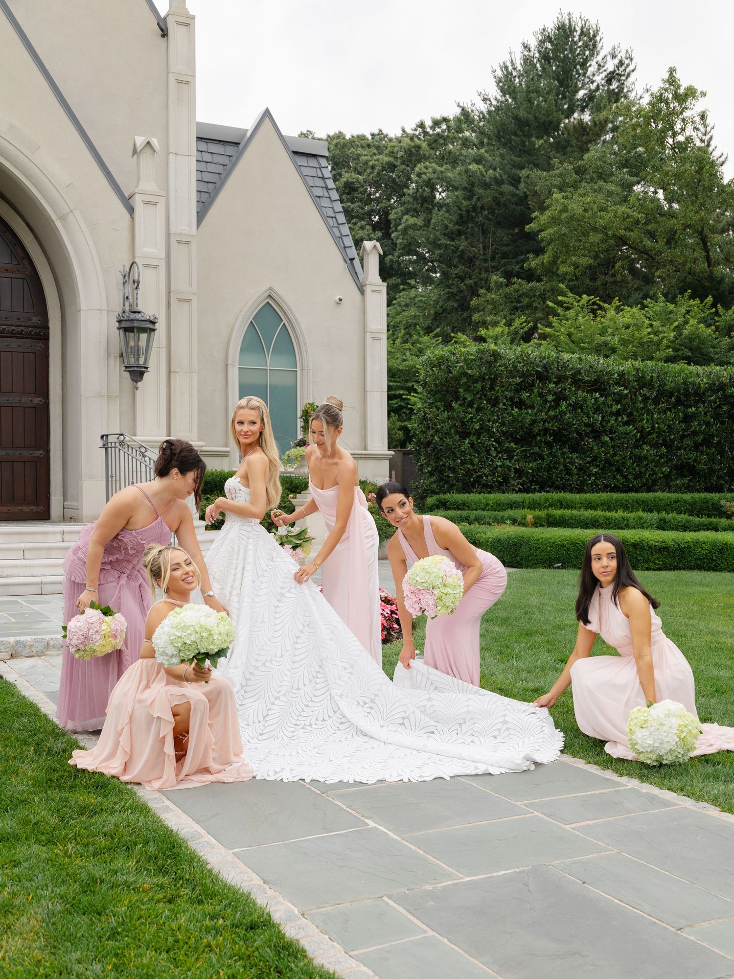 Bridesmaids in pink dresses arrange bride's wedding gown train outside a white stone church on a sunny day.
