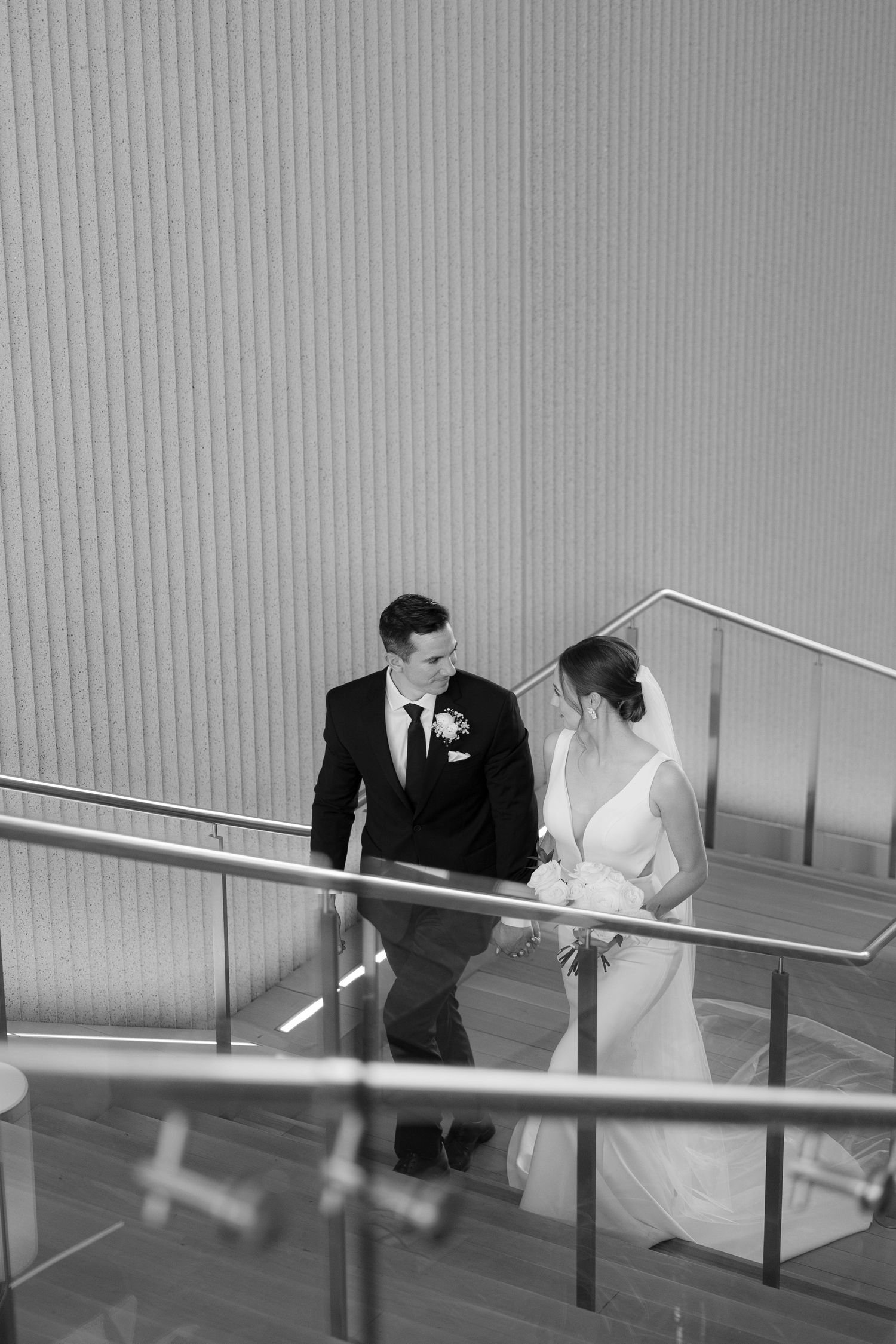 Black and white photo sequence shows a couple in wedding attire descending a modern staircase together.