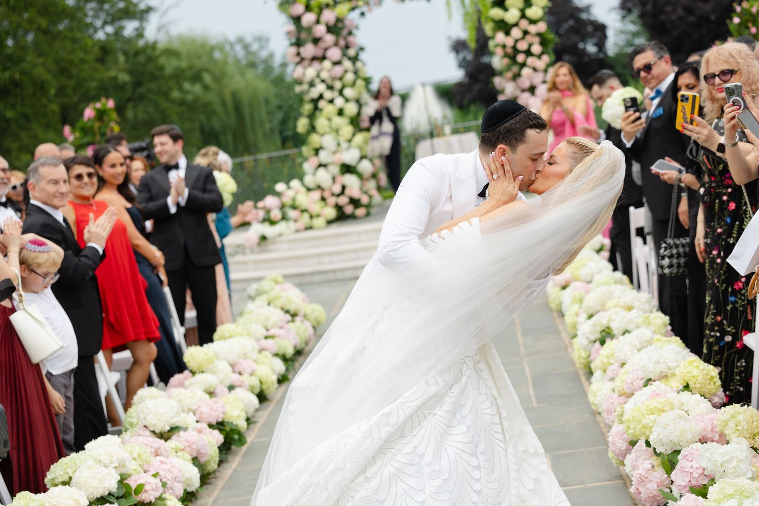 Wedding guests shower newlyweds with confetti as they walk along a flower-lined path during outdoor celebration.