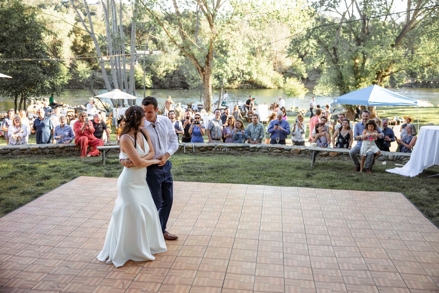 A couple shares their first dance on an outdoor patio while wedding guests watch from the lawn on a sunny afternoon.