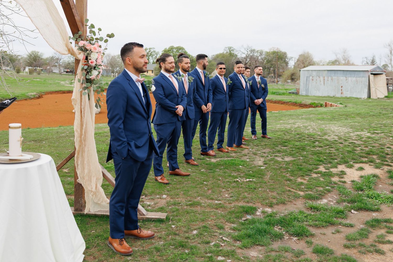 A wedding party in navy blue suits stands in a line outdoors on a grassy field during a ceremony.