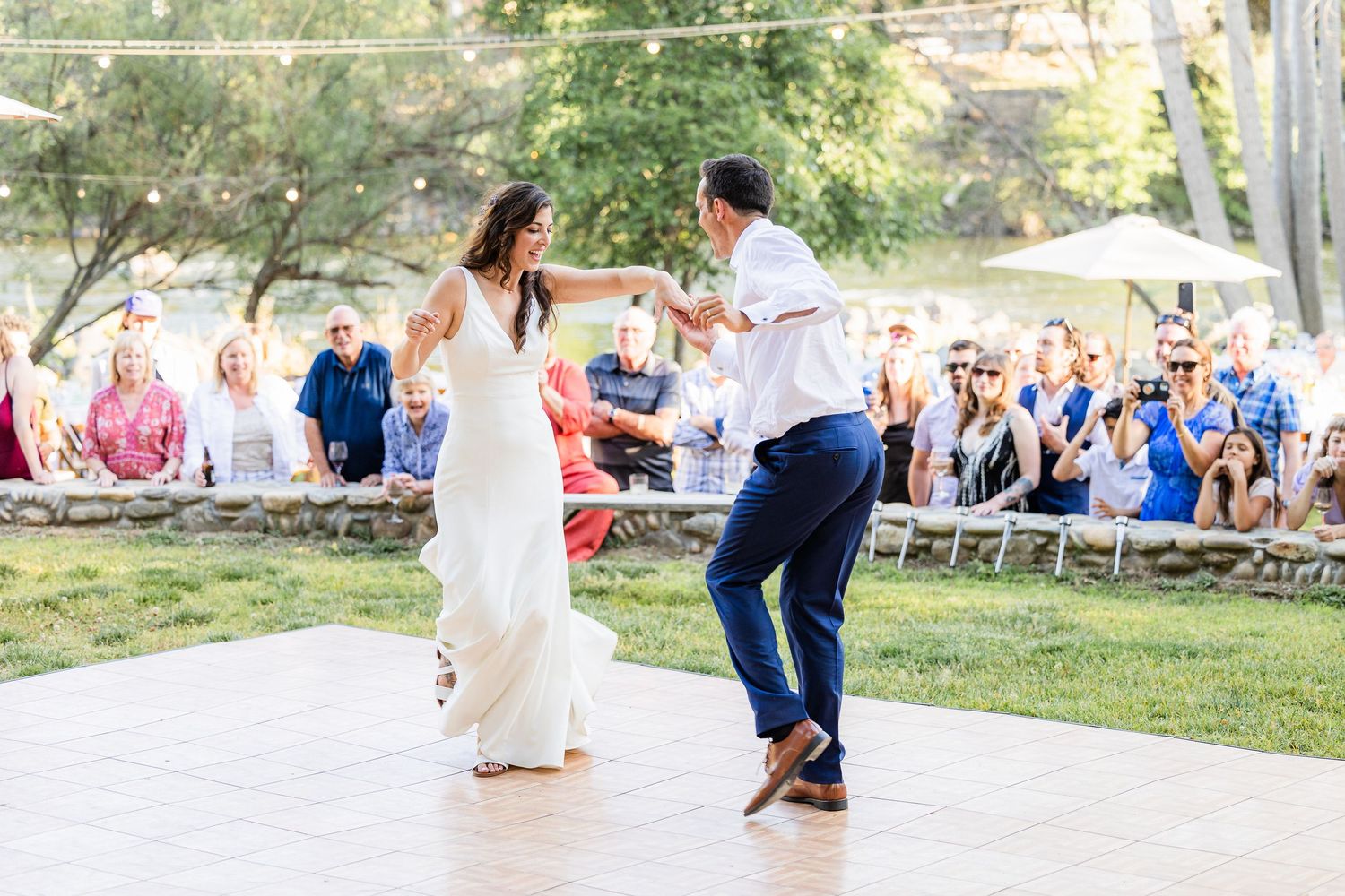 A couple dances together in an outdoor park setting surrounded by trees and other people enjoying the afternoon.