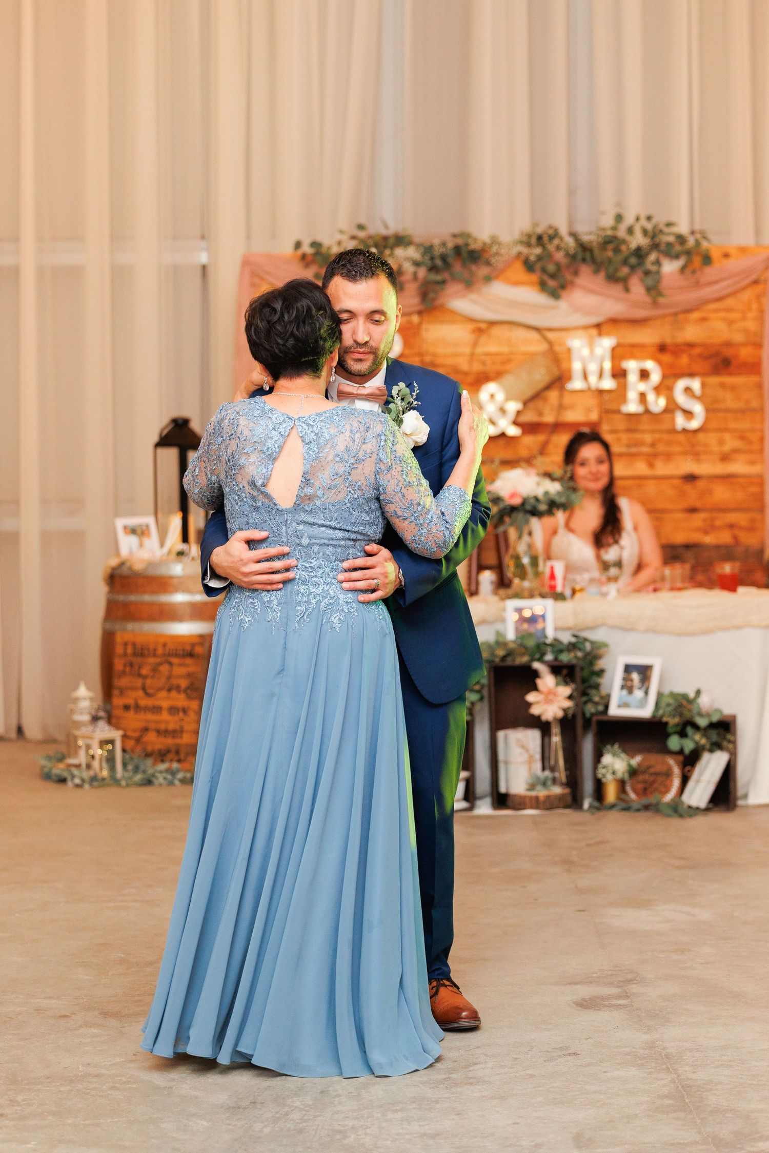 A couple shares a dance at their wedding reception with rustic wooden decor and 'MRS' signage in the background.