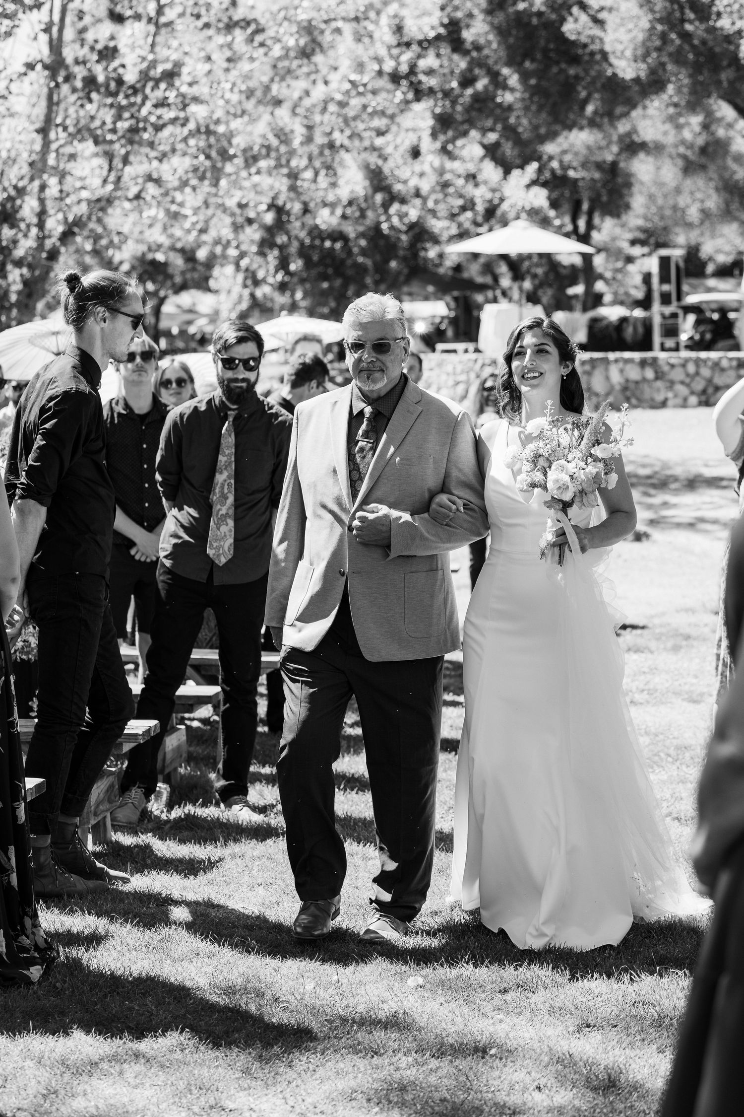 Black and white photo of an outdoor wedding ceremony with a bride in a white dress walking down the aisle with an escort.