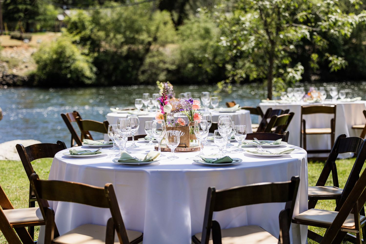 An elegant outdoor wedding reception table with white linens and place settings beside a river in a garden setting.