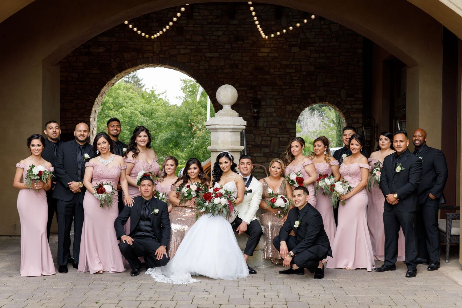 Wedding party in pink bridesmaid dresses and dark suits pose together under stone archway for formal photos.