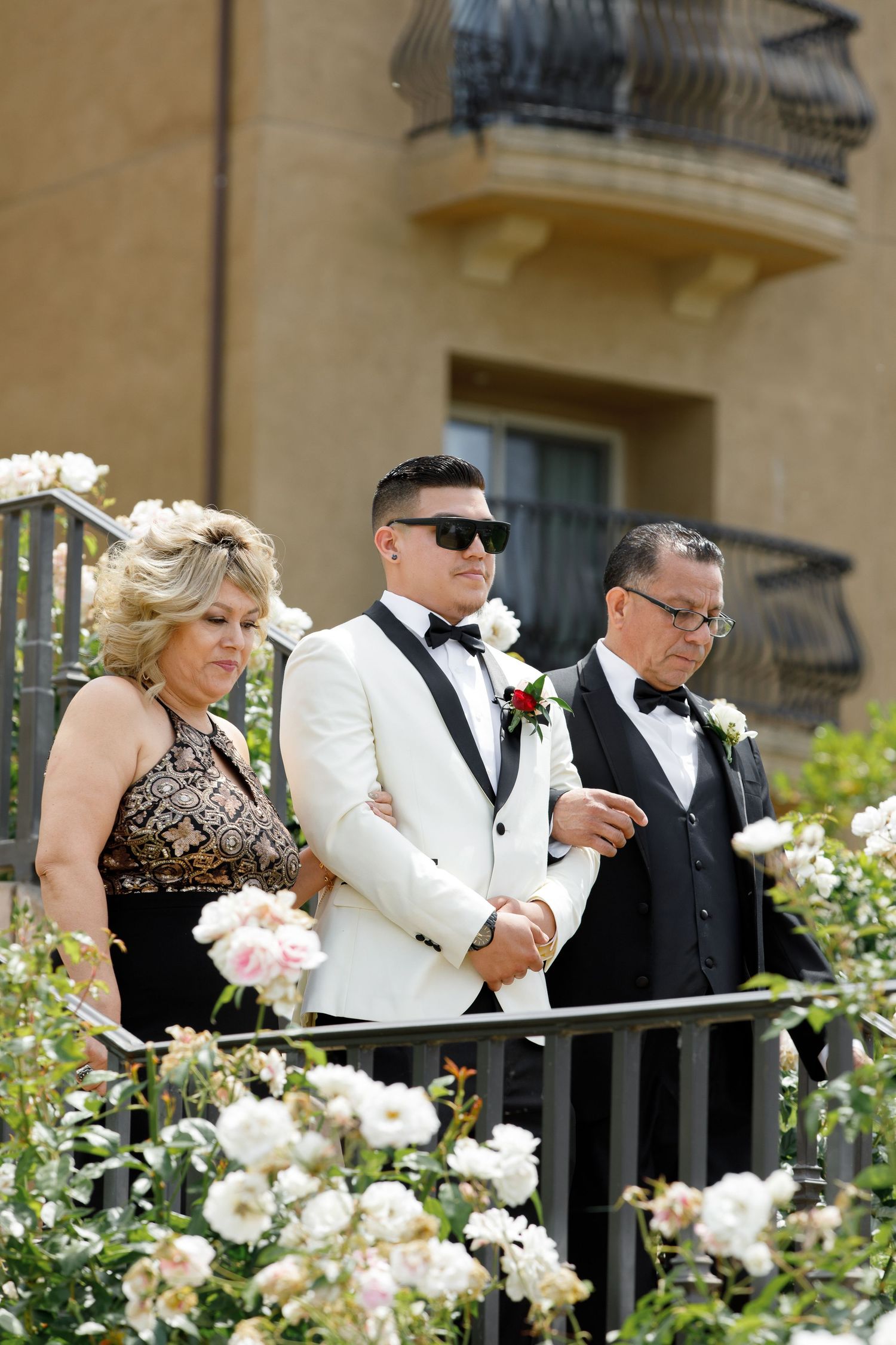 Formally dressed wedding guests in black and white attire stand along a railing adorned with pink and white roses.