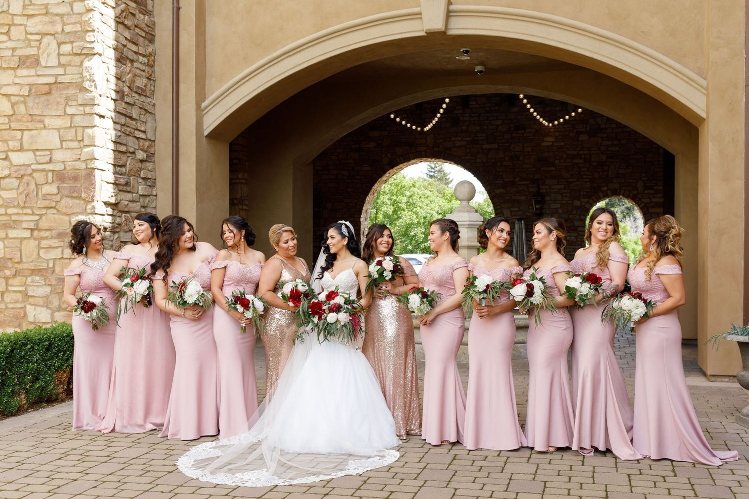 A wedding party wearing pink and white dresses poses together in an elegant stone archway setting.