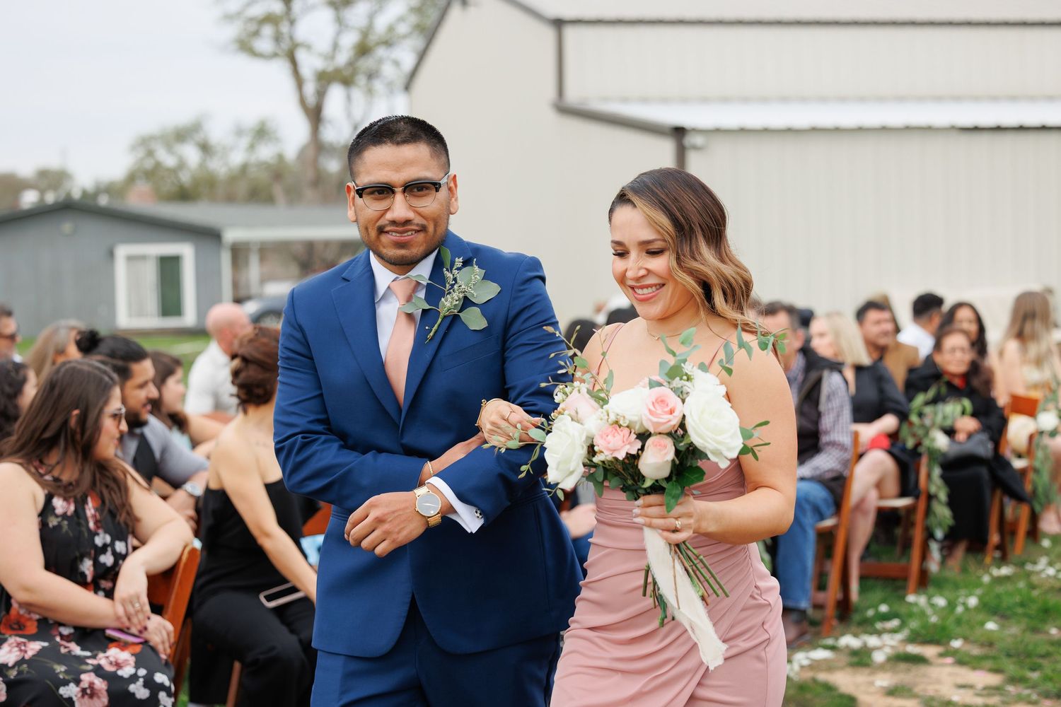 Wedding party members dance and celebrate together at an outdoor reception venue with white walls and greenery.