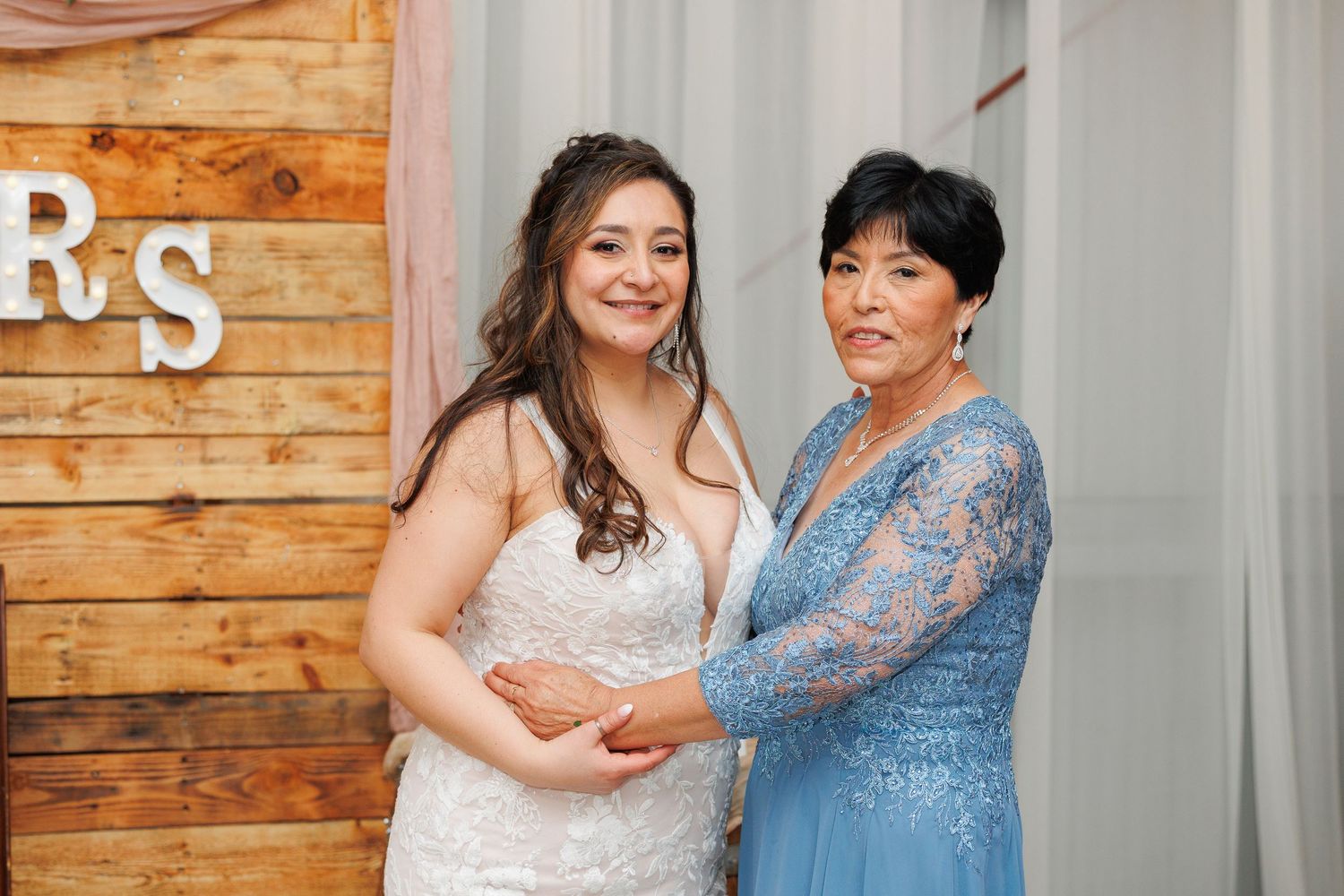 A bride in a white dress shares a joyful moment with someone wearing a blue lace dress at an indoor venue.