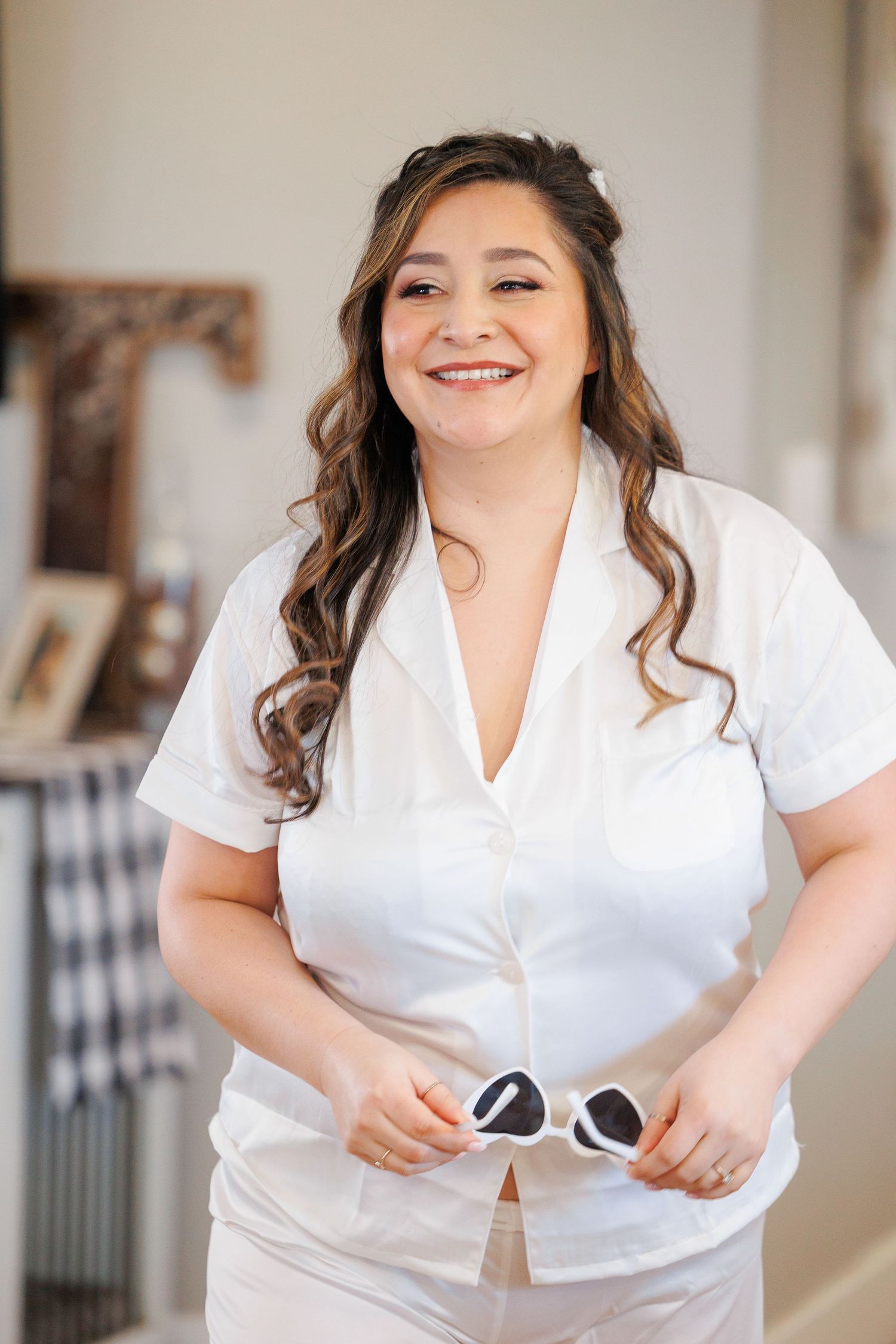 Smiling woman in white outfit holds sunglasses while posing indoors with long wavy brown hair.