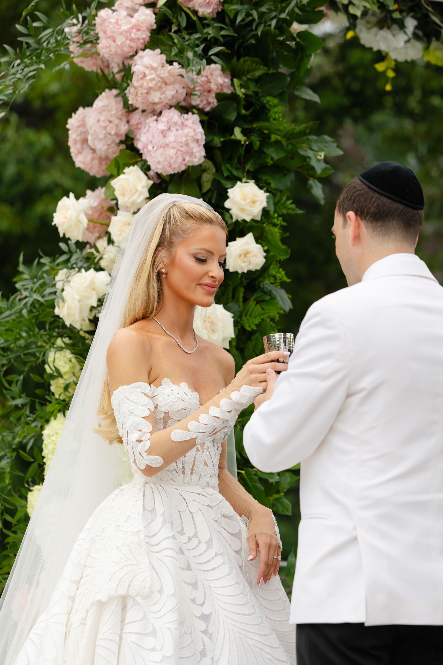 Bride and groom sharing a cup during an outdoor Jewish wedding ceremony surrounded by pink and white floral arch.