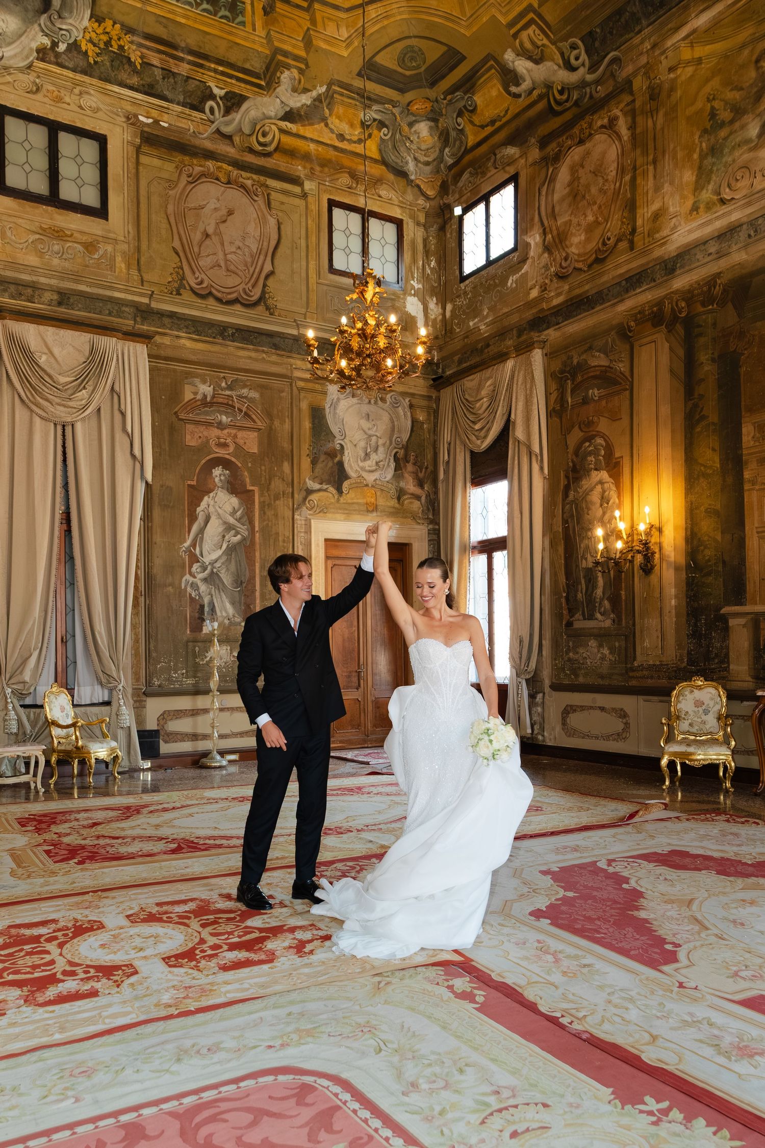 A couple dances in an ornate baroque palace ballroom with gilded walls, crystal chandeliers and a red patterned carpet.