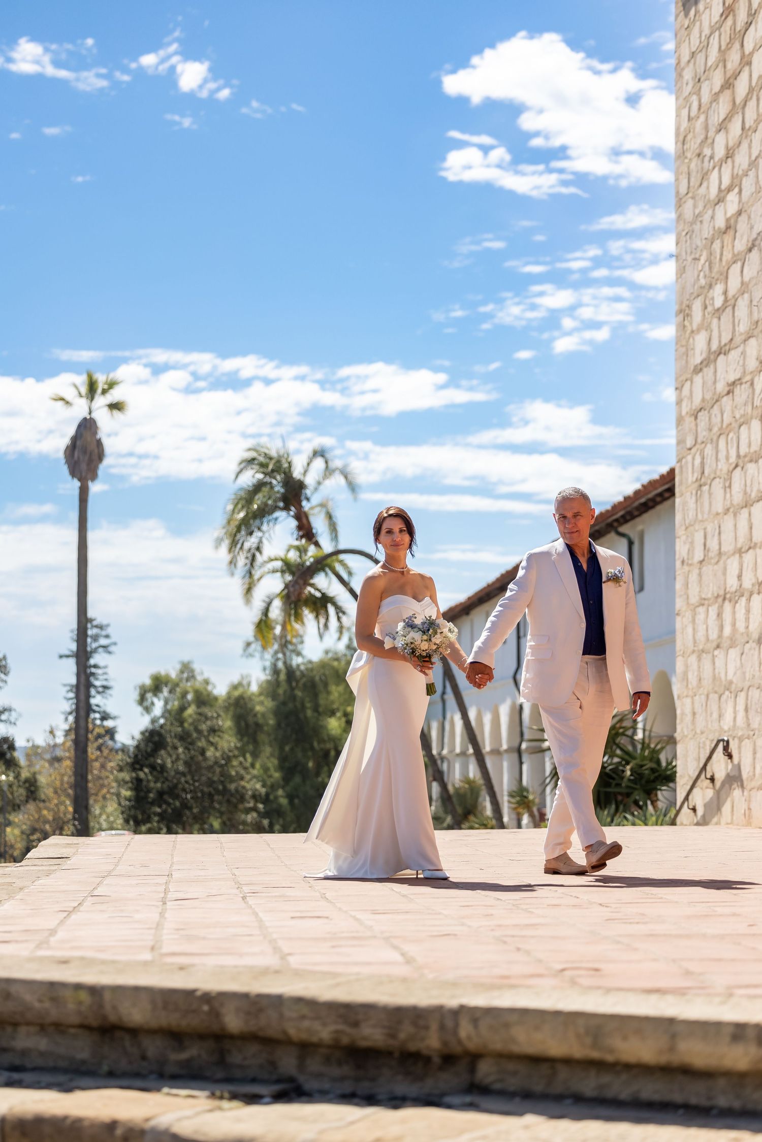 Wedding couple poses against stone wall backdrop with palm trees and blue sky.