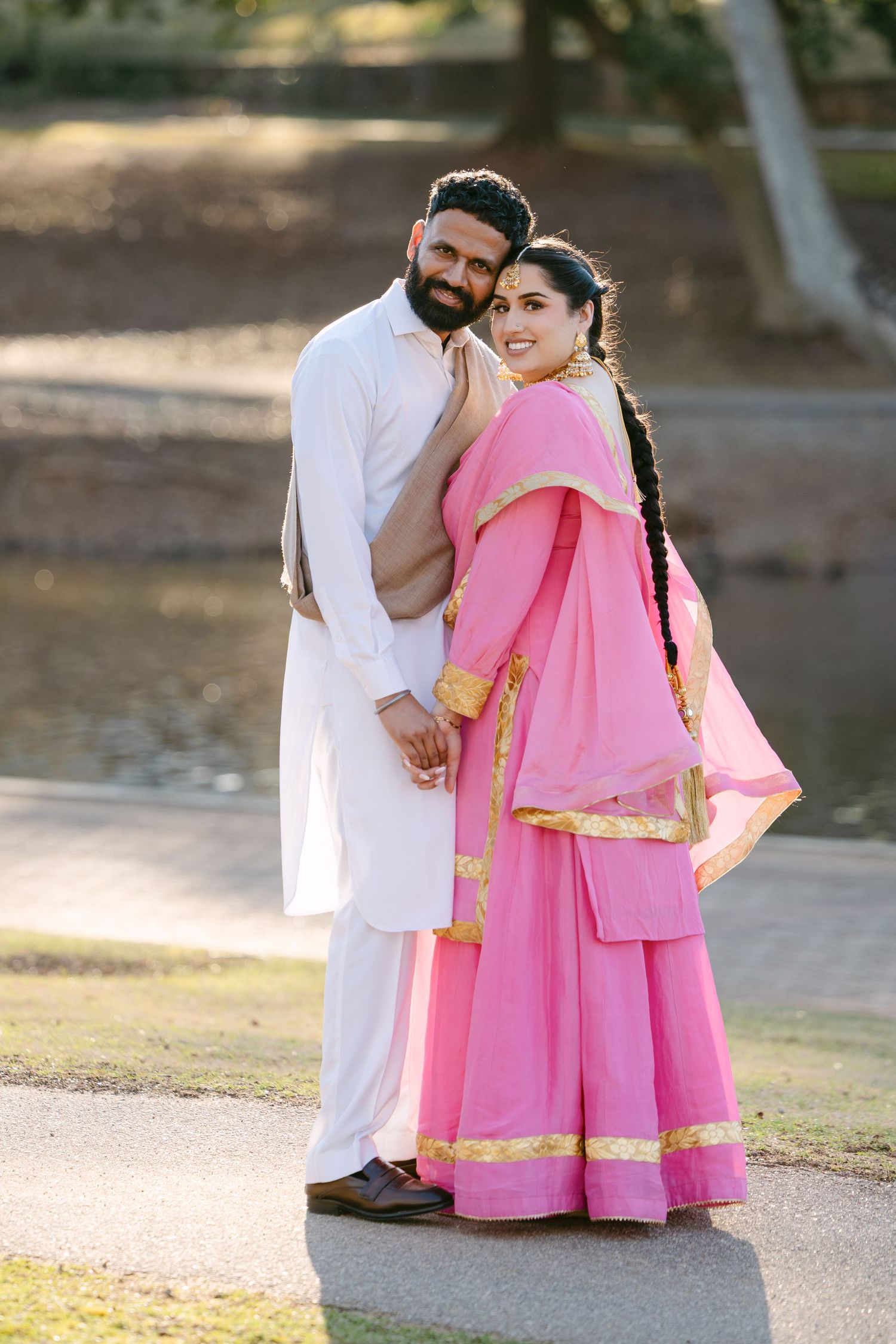 A couple in traditional attire shares tender moments outdoors, with the woman wearing a flowing pink dress with gold trim.