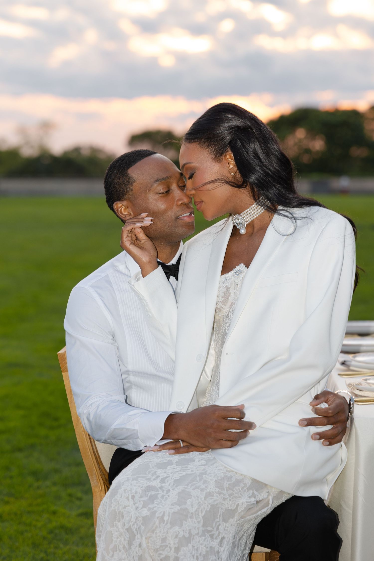 A romantic wedding couple shares an intimate moment outdoors on a grassy field at sunset.
