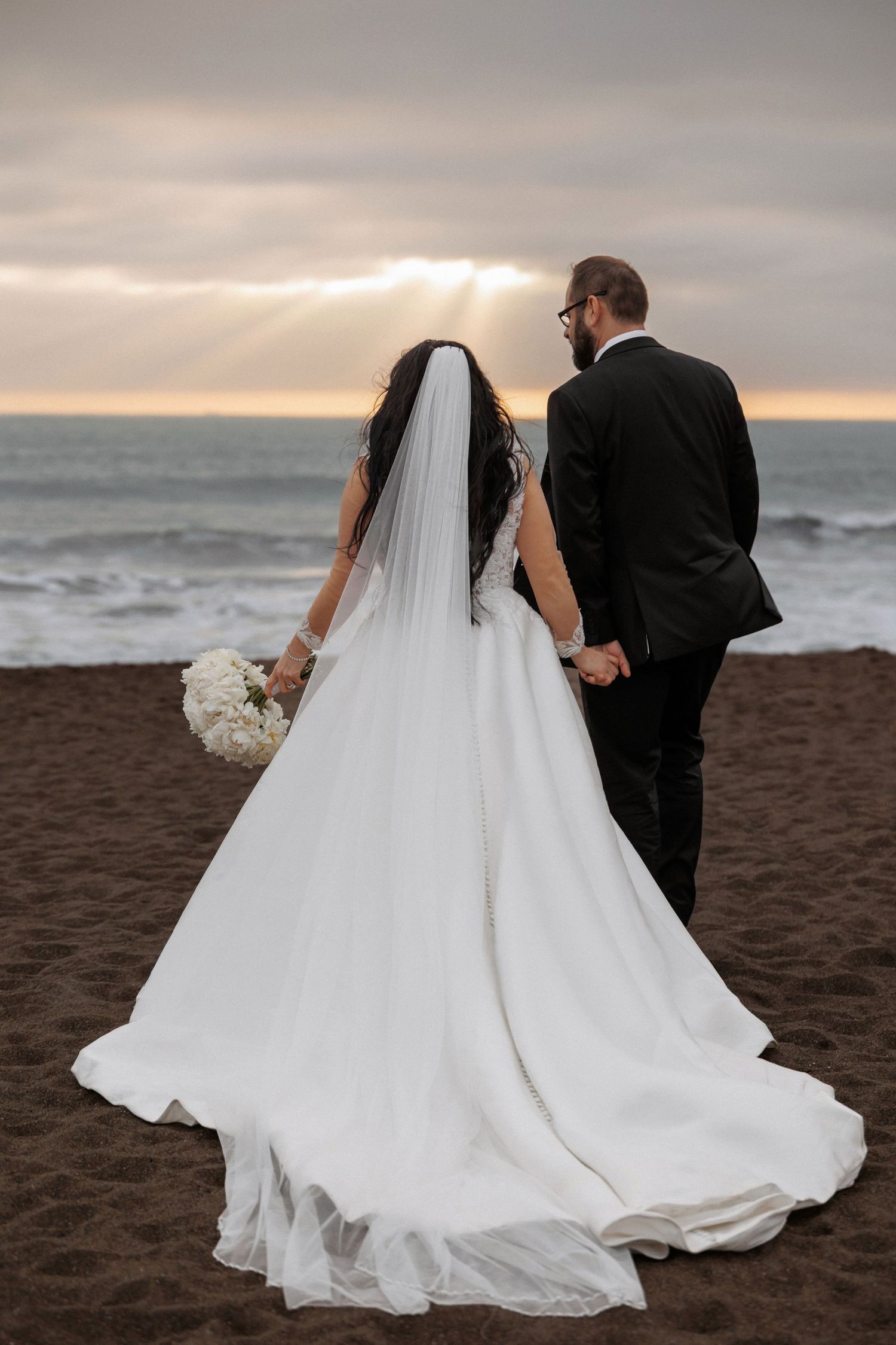 A couple in wedding attire walk along a misty beach at sunset with the bride's flowing white dress trailing behind.