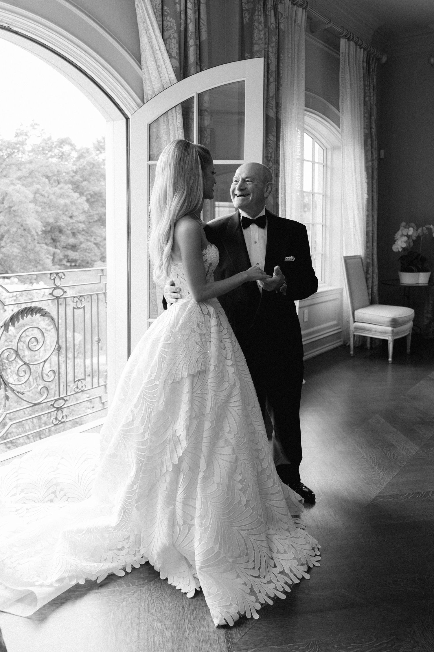 Black and white photo of a bride in a lace gown sharing a joyful dance with an older man in a tuxedo.
