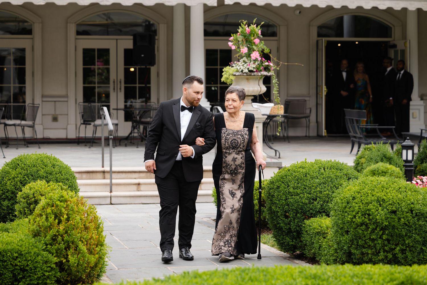 Elegant wedding party walks along a landscaped pathway with manicured hedges and white columns of a grand building.