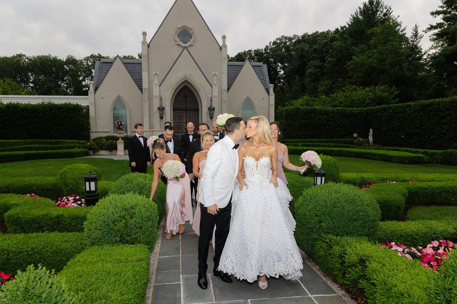 Wedding party walks through a lush garden path beside a white church on an overcast day.