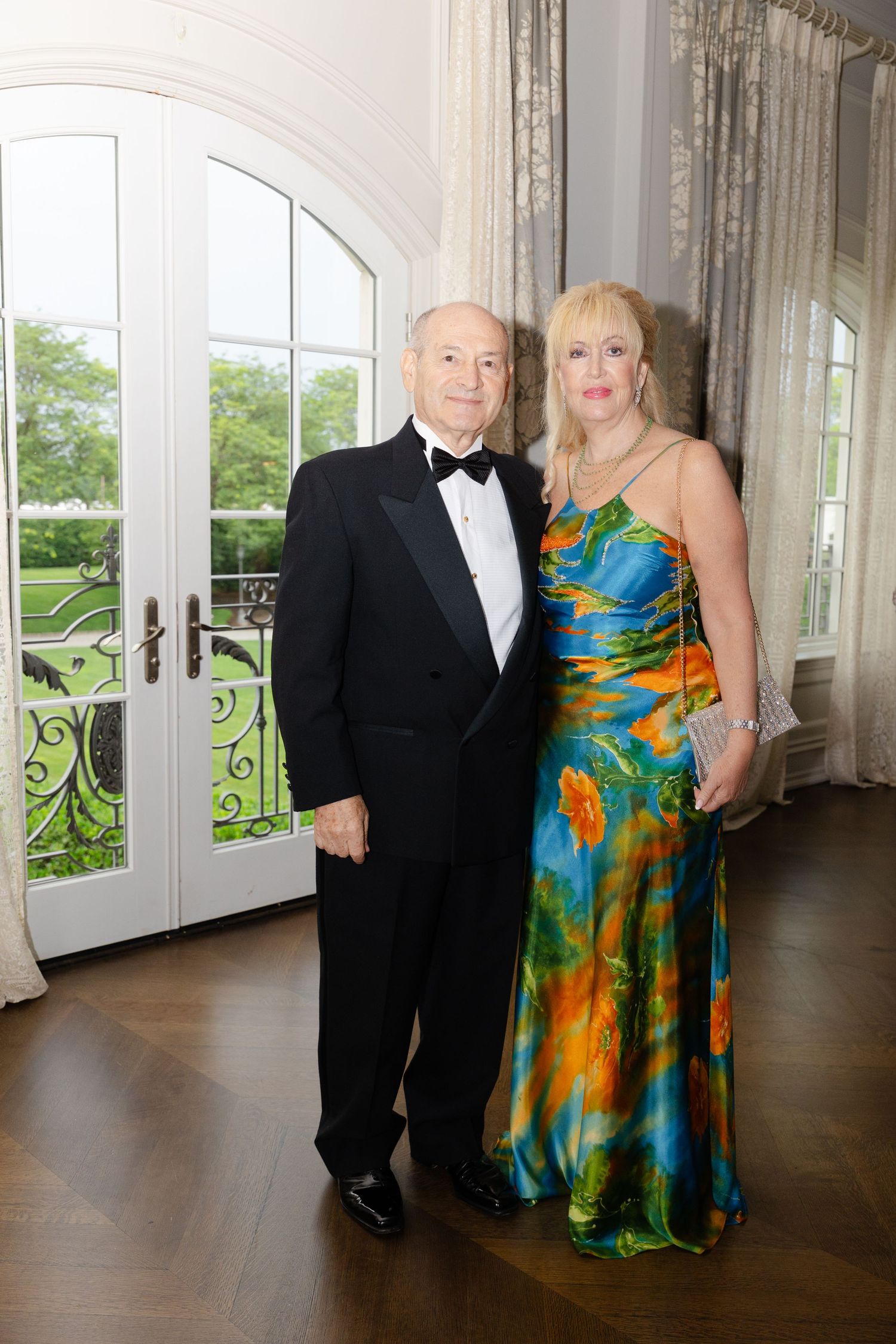 A couple in formal attire poses in an elegant hallway with French doors and natural light streaming in.