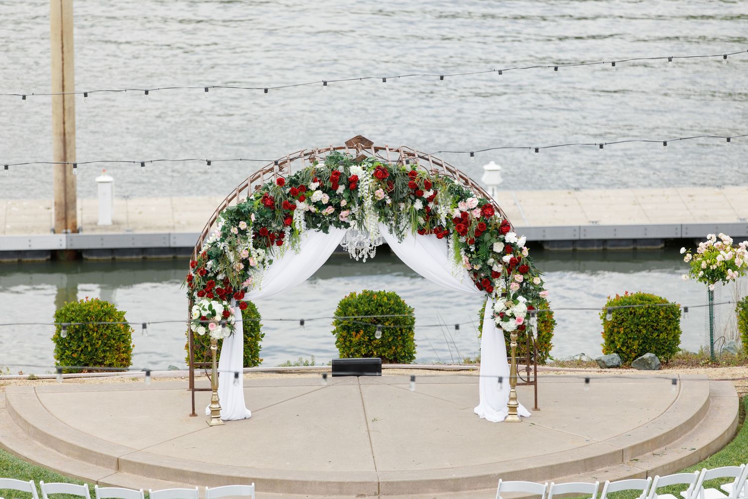 White wedding arch decorated with red roses and greenery stands against a rustic whitewashed brick wall.