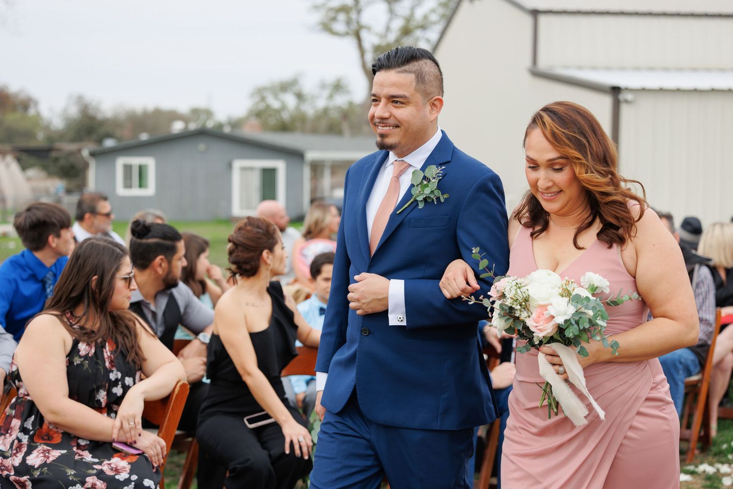 Wedding party in navy suits and blush pink dresses celebrates joyfully during outdoor reception.