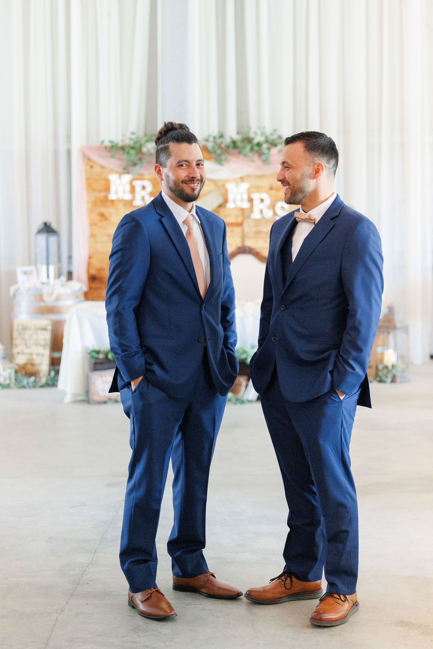 Two people in navy blue suits share a laugh while standing in an elegant event space with rustic decor.