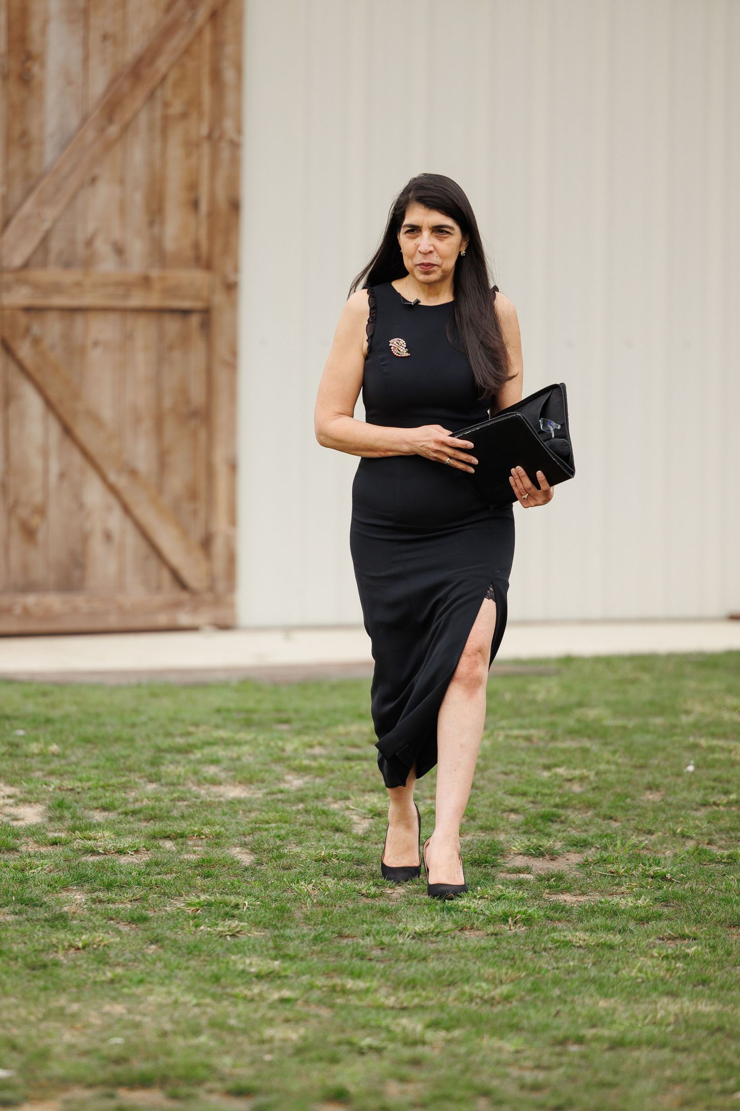 A person in an elegant black dress with a side slit walks on grass near a rustic wooden door.