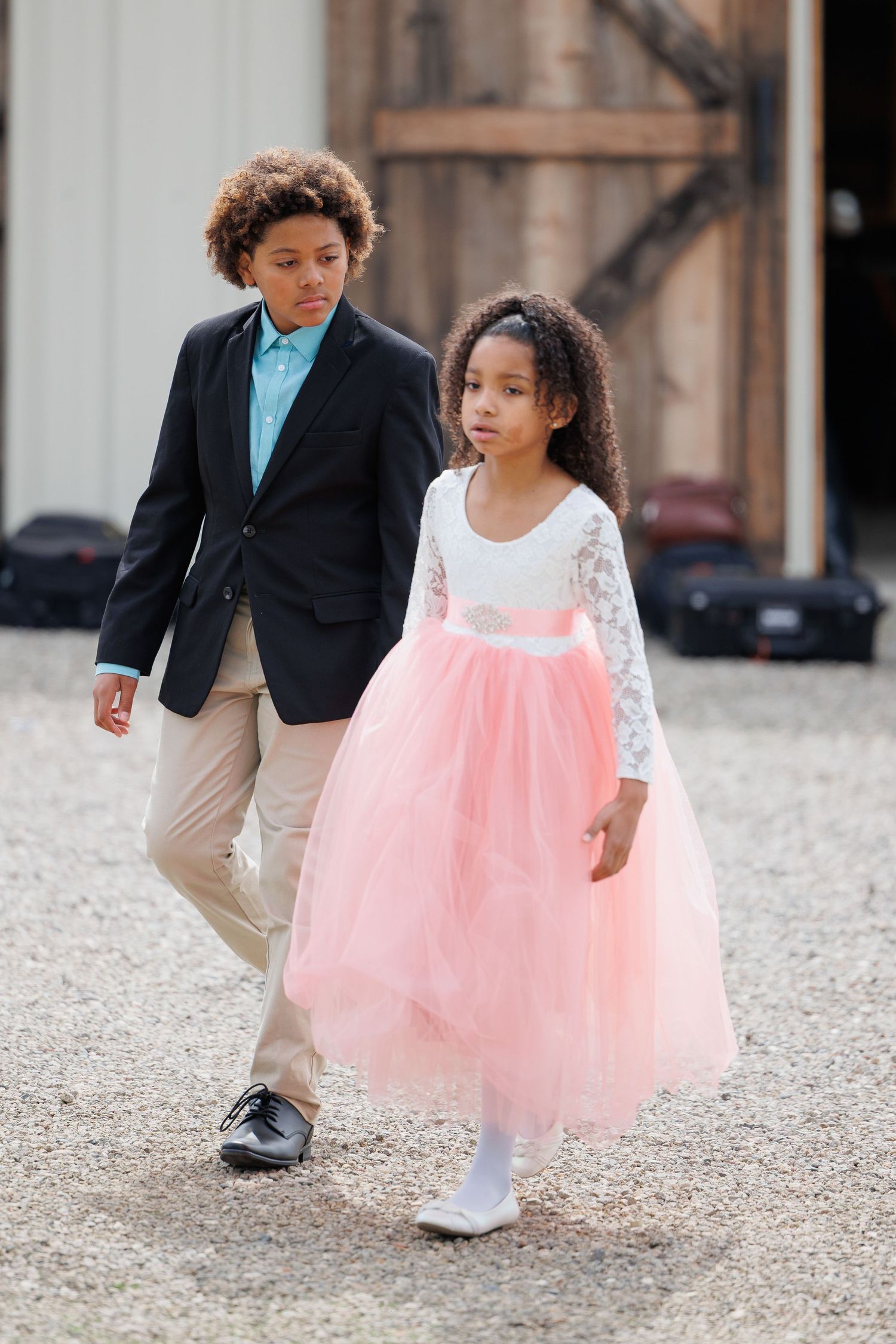 Two children in formal attire walk together on a gravel path, with one wearing a pink tulle dress.