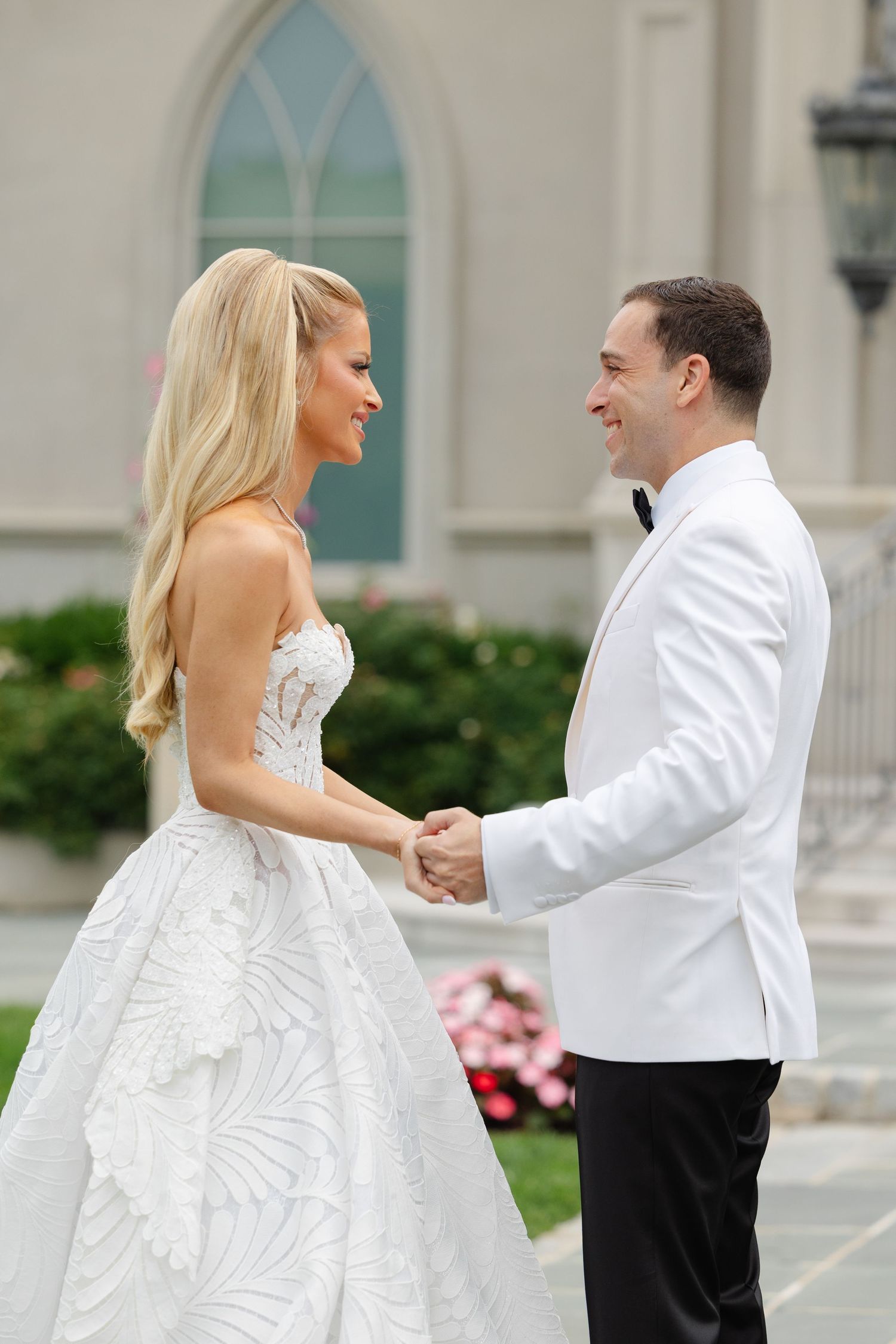 A bride and groom share a joyful moment outside a church with an arched window in the background.