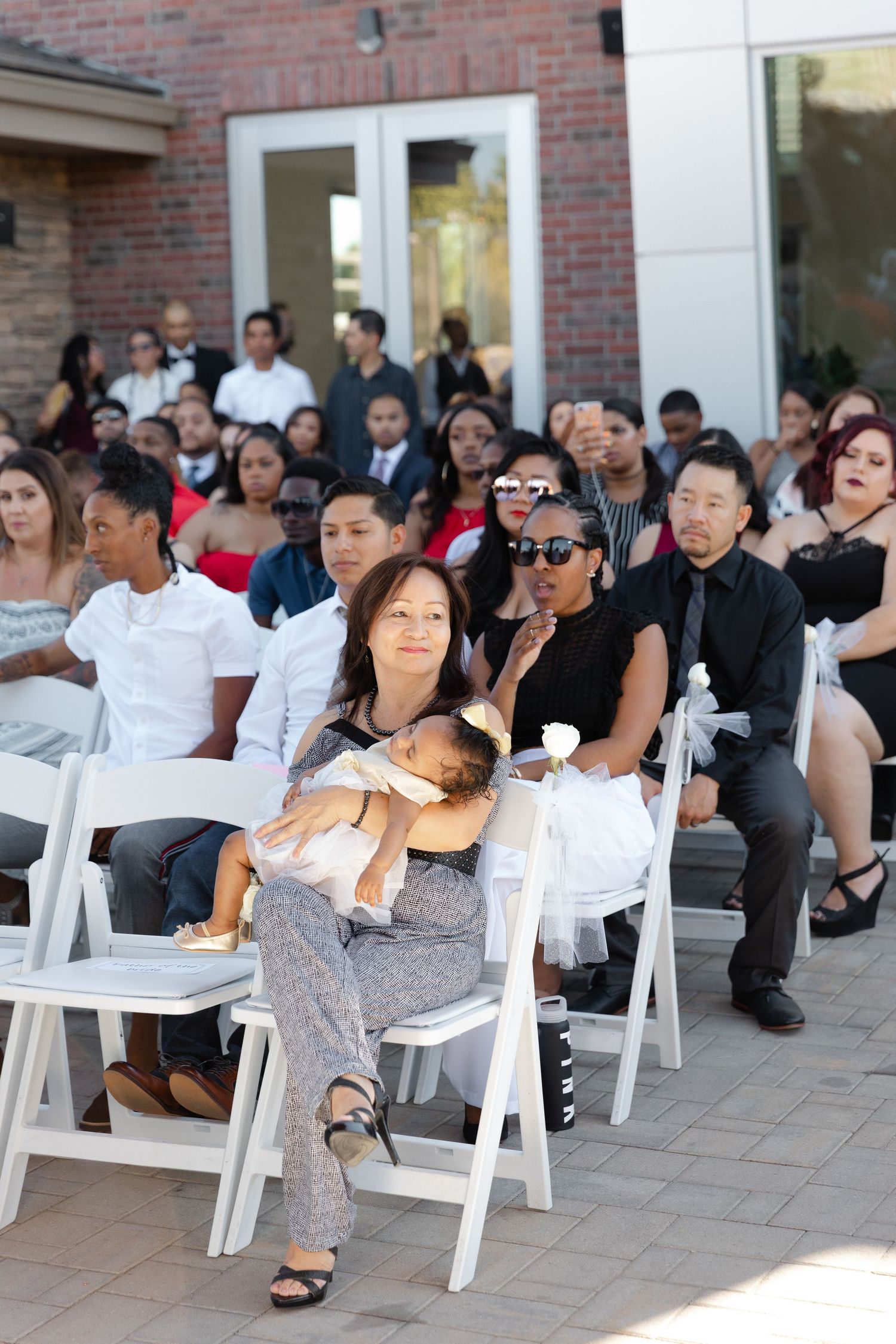 A diverse group of guests seated in white chairs at an outdoor event near a brick building.