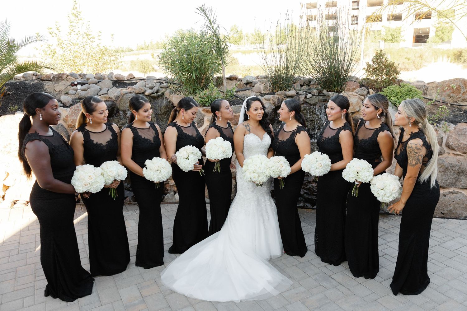 A bride in white stands with her bridesmaids wearing elegant black dresses holding white bouquets at an outdoor venue.