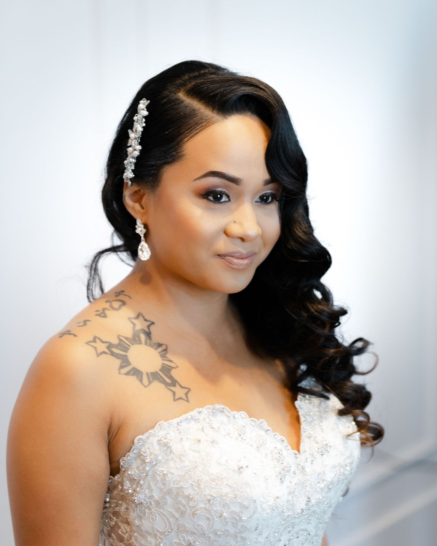 Bride in white beaded strapless wedding gown with curled black hair and floral neck tattoo poses against light background.