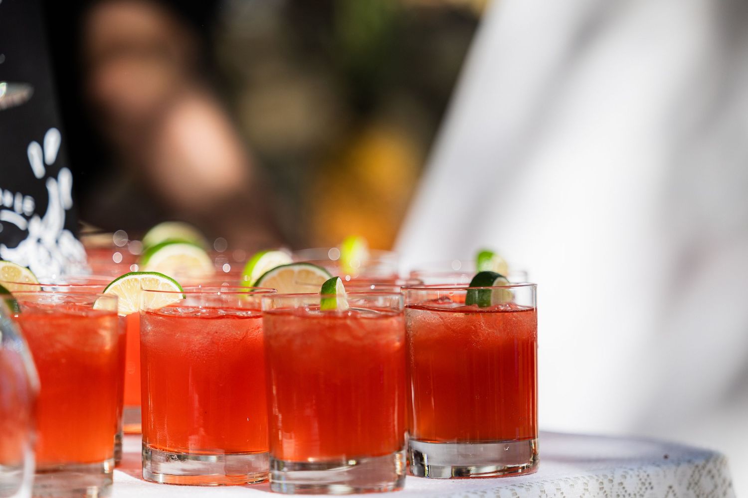 A row of red cocktails garnished with lime wedges sits on a bar counter in a dimly lit setting.