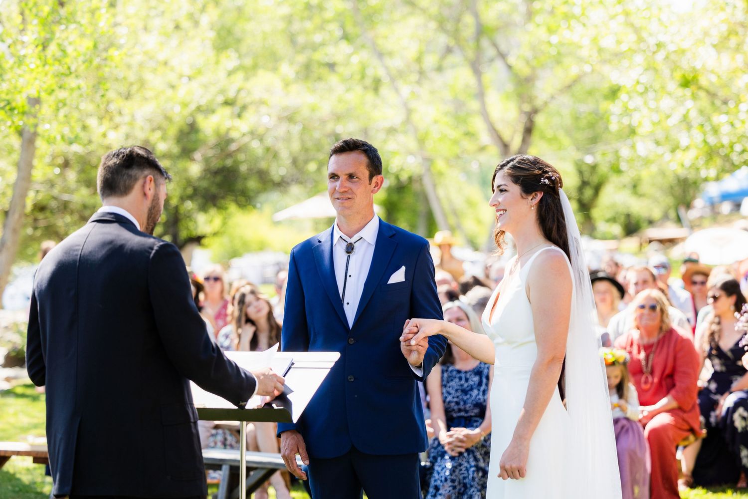 Wedding guests gather outdoors under sunlit trees during a summer celebration ceremony.