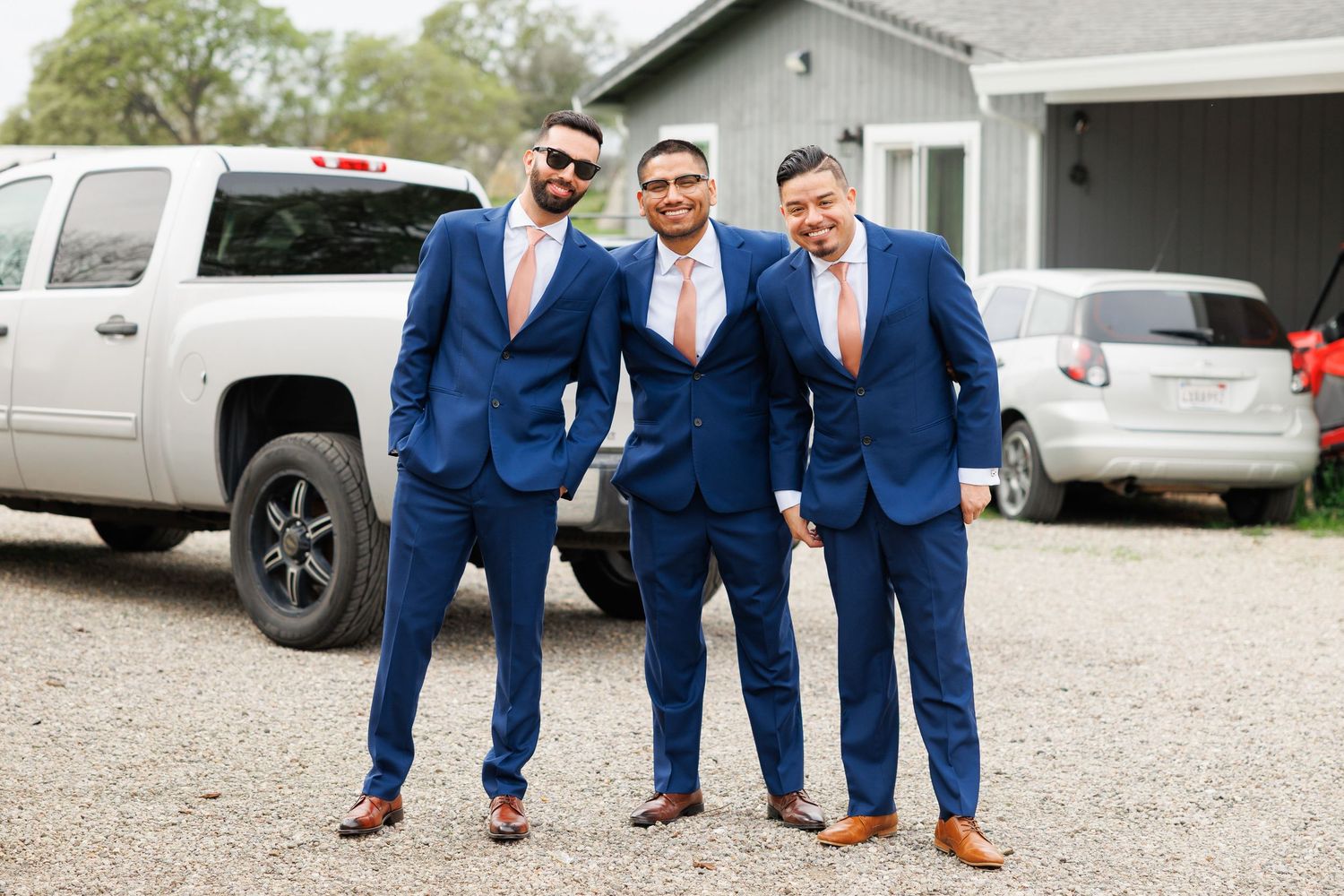 A group of groomsmen in matching navy blue suits pose playfully in a gravel parking lot.