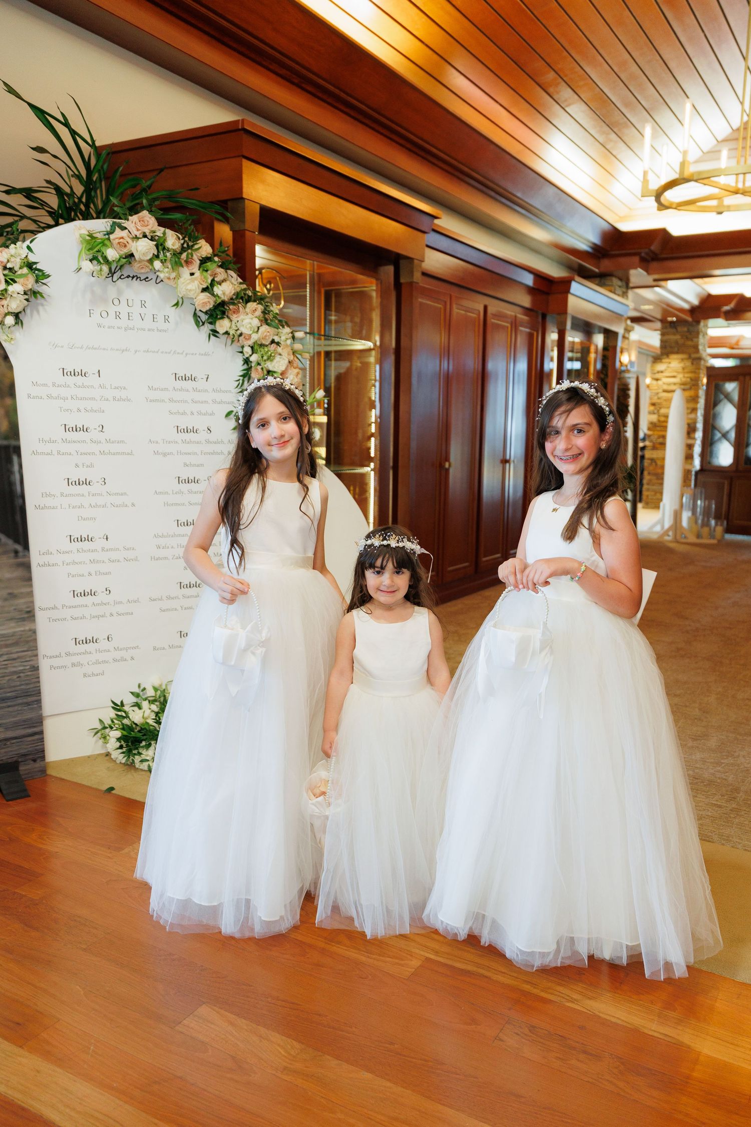 Three flower girls in white tulle dresses pose playfully in a wood-paneled hallway with hardwood floors.