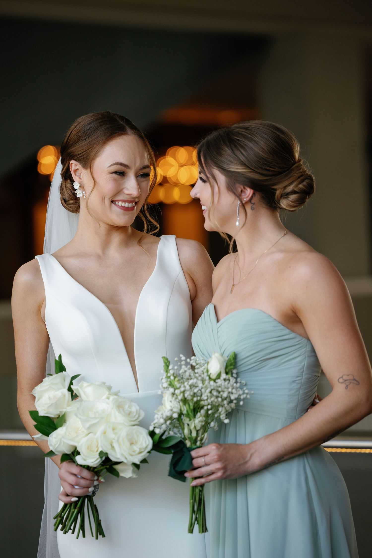 Two women in formal dresses share a joyful moment while holding white floral bouquets against a warm bokeh background.