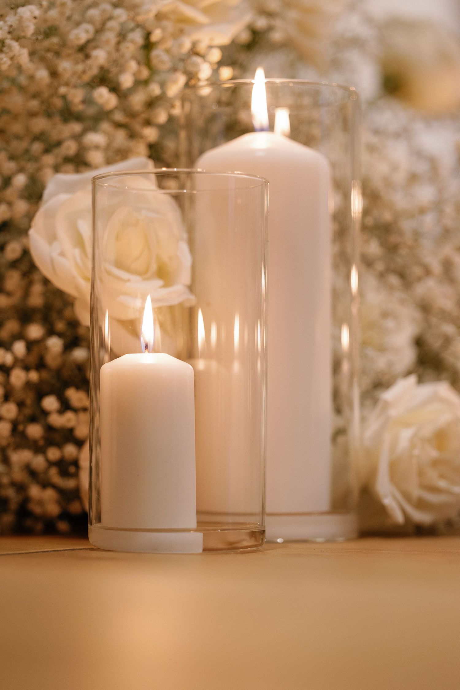 Elegant white pillar candles in glass cylinders glow softly beside delicate white roses and baby's breath flowers.
