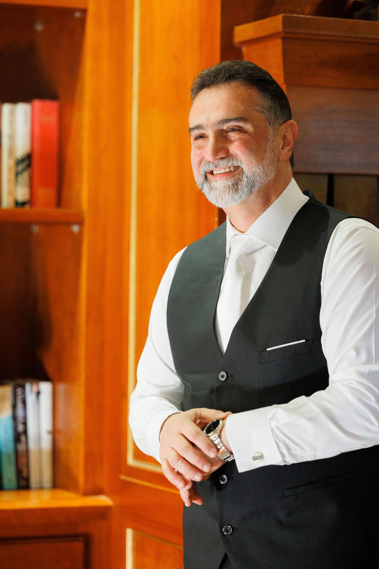 A person in formal attire adjusts their vest while standing in a warmly lit room with wooden paneling.