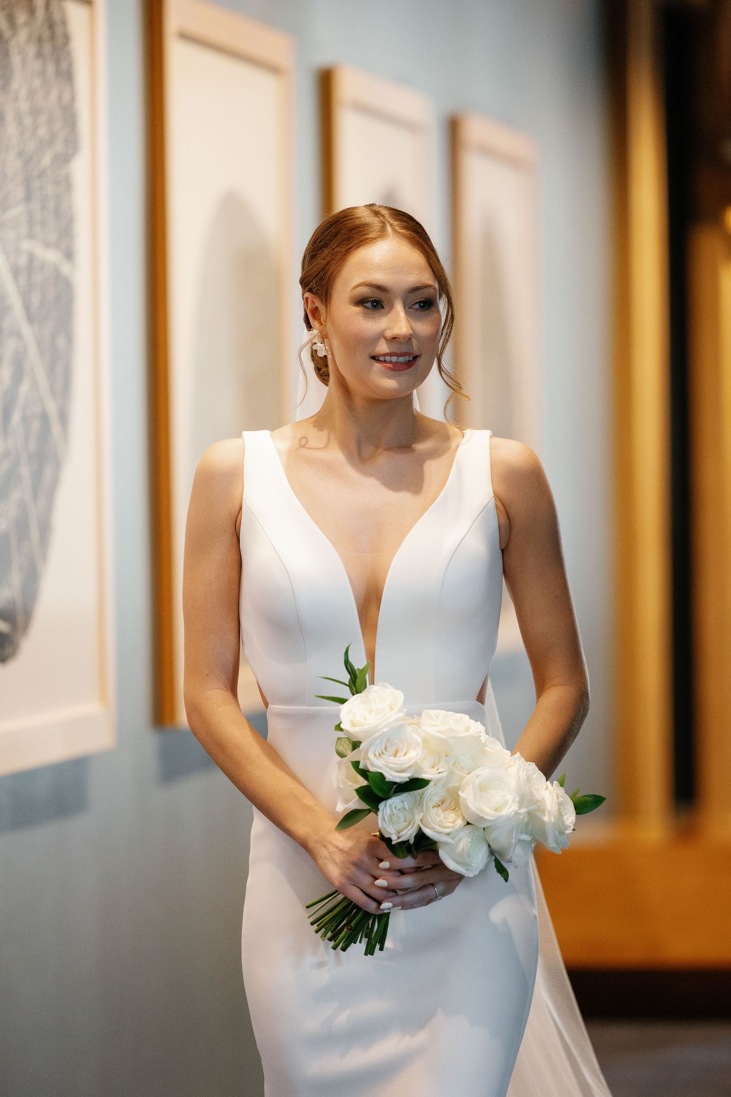 A bride in an elegant white v-neck wedding dress holds a bouquet of white roses in a hotel hallway.