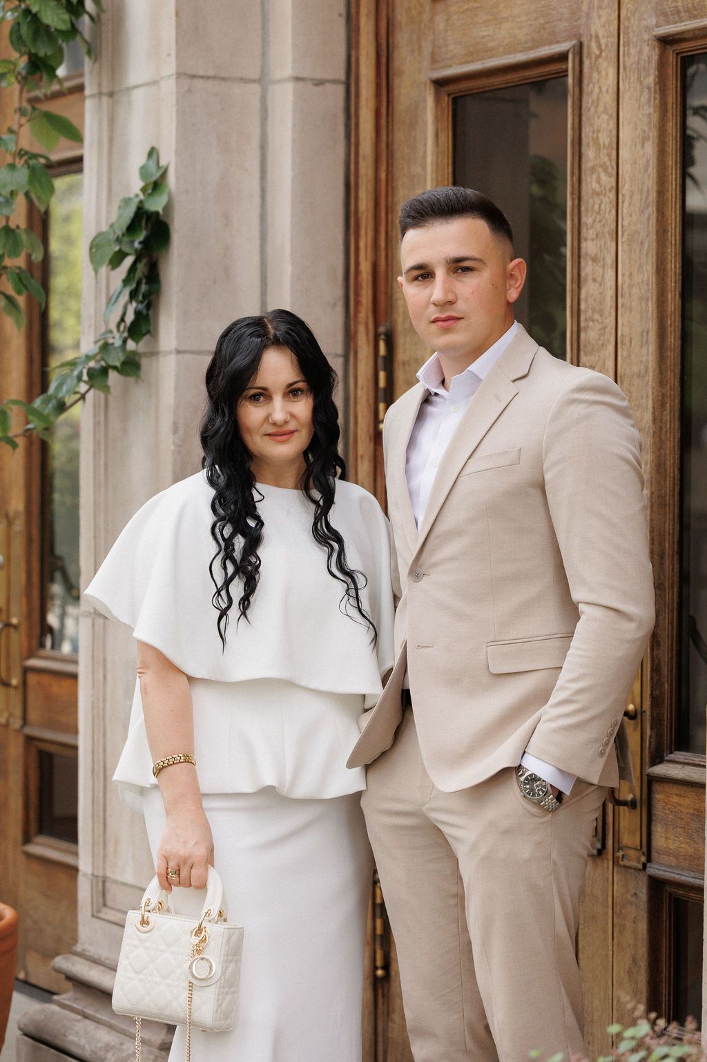 A couple in elegant wedding attire poses against a stone building with ivy, wearing beige suit and white dress.