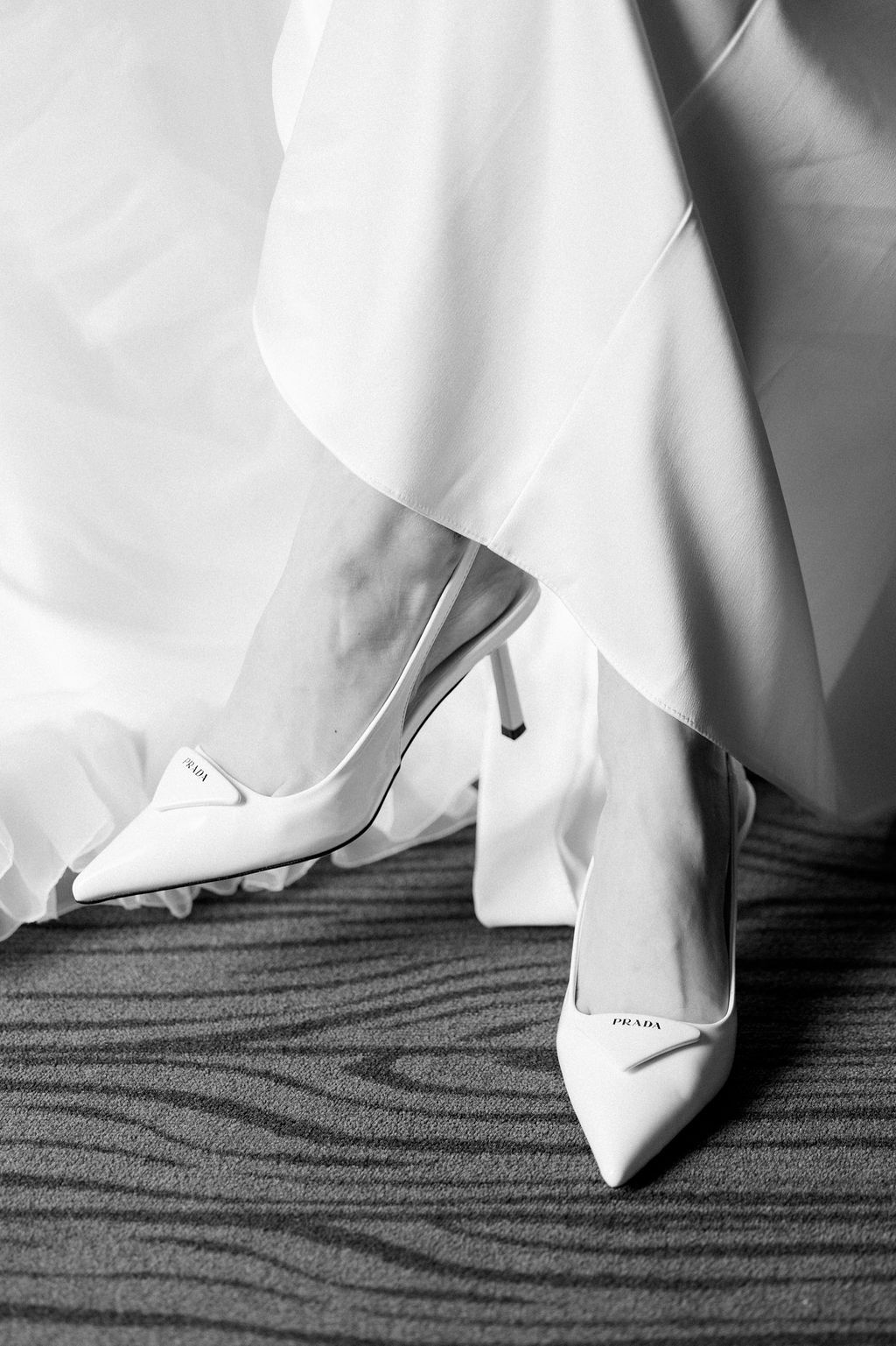 Close-up of white pointed-toe heels and wedding dress hem on textured carpet.