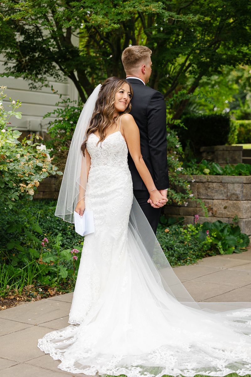A couple in wedding attire holds hands while walking along a stone path surrounded by lush greenery.